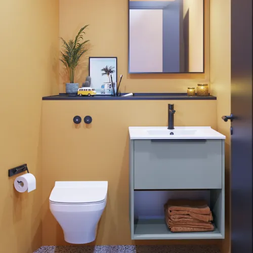 Pienen kylpyhuoneen inspiraatio vinokattoisilla ja lämpimän keltaisilla seinillä, tyylikäs vaaleansininen pesuallaskaappi, ohut ja tyylikäs pesuallas sekä mustareunainen neliönmuotoinen peili ja käytännöllinen lamppu.